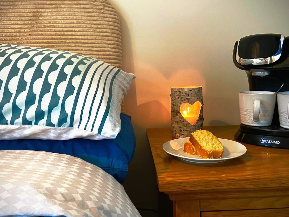 A bedside table holds a heart-shaped candle within a bark candle holder, alongside a plate with a slice of cake. A Tassimo coffee machine and stacked cups are visible, with neatly arranged cushions and a cozy bed in the background.