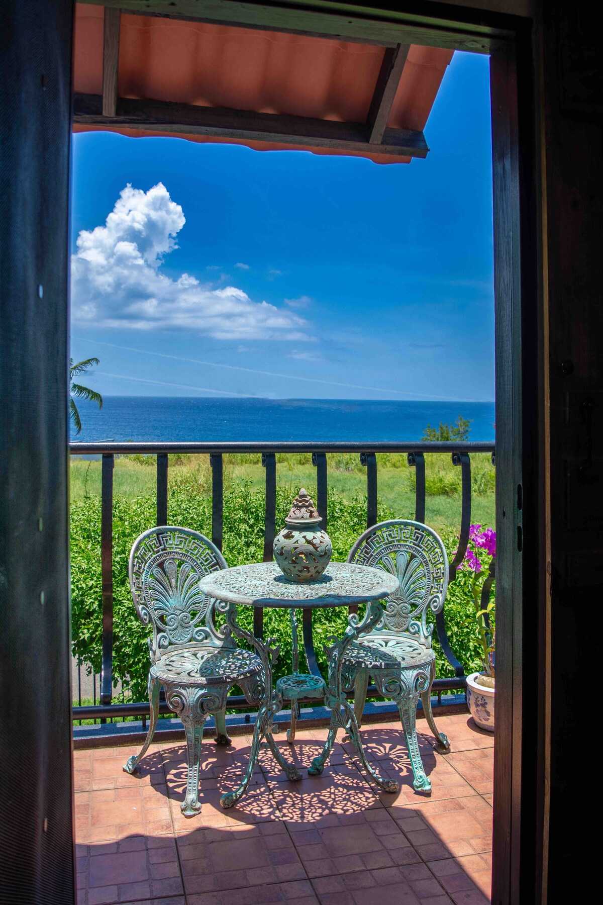 A small balcony is visible through an open door, featuring a round table and two decorative chairs made of wrought iron. A ceramic vase sits at the center of the table. In the background, a view of the sea and a bright blue sky is presented.