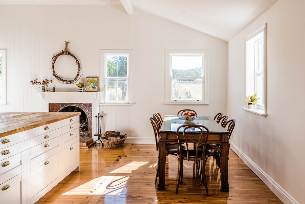A bright, open kitchen and dining area is illuminated by natural light streaming through two windows. A rustic wooden table surrounded by six chairs sits centrally, complemented by a vintage fireplace and decorative wall wreath, enhancing the space's charm.