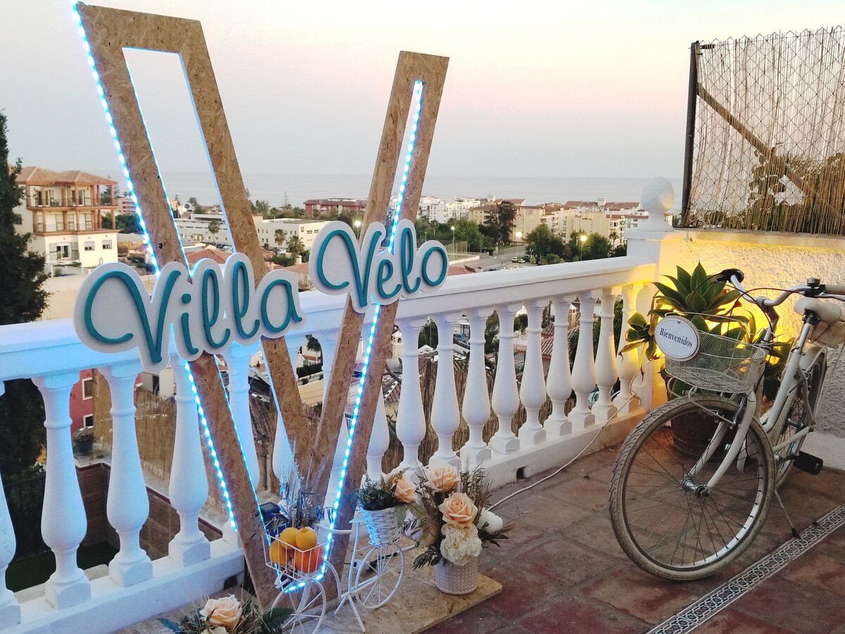 An outdoor terrace features a decorative sign with the name 'Villa Velo' illuminated by soft lights. A vintage bicycle is parked nearby, complemented by a small planter of lush greenery and ornamental flowers, with a serene ocean view in the background.