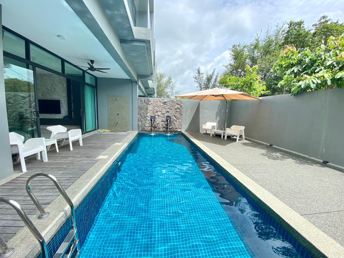 A private pool area is framed by a modern design, featuring a clear blue pool complemented by a sunshade and lounge chairs. Large glass doors create opportunities for natural light, while lush greenery enhances the serene environment.