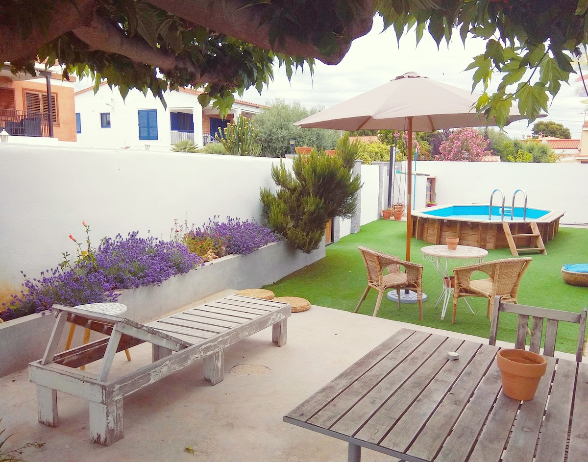 An outdoor garden area is featured, equipped with a wooden dining table and chairs. A lounge chair rests beside a small flower bed with blooming lavender. An umbrella provides shade over the pool area, which includes a swimming pool surrounded by artificial grass.