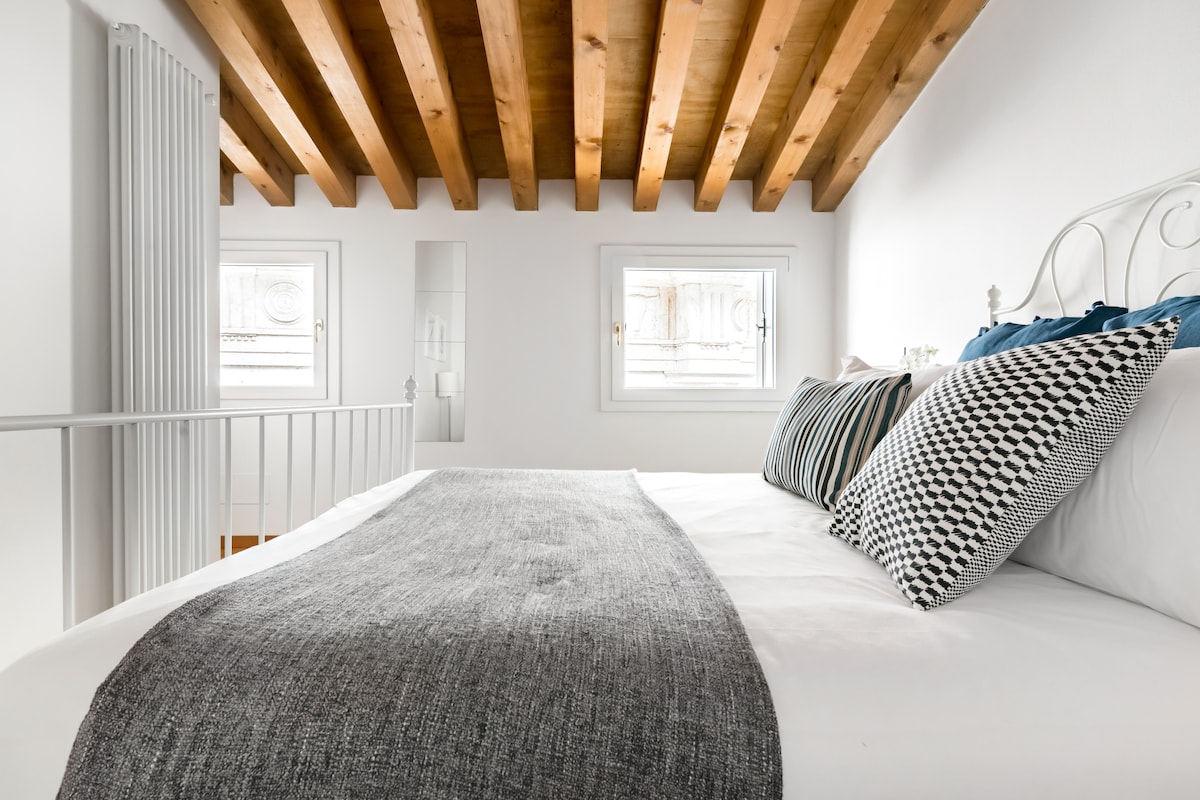 A serene bedroom is presented with a spacious bed featuring a neutral-colored blanket. The wood-beam ceiling adds warmth, while natural light enters through two windows. Decorative pillows in varied patterns enhance the inviting feel of the room.
