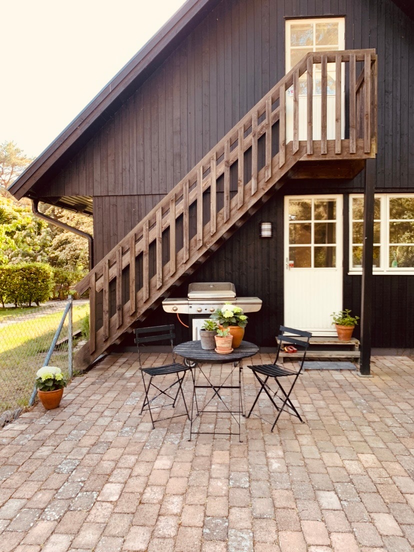 An outdoor patio area features a circular table surrounded by two black chairs, complemented by potted plants. A barbecue grill is positioned nearby, and an exterior wooden staircase leads to the upper level of the two-story building.