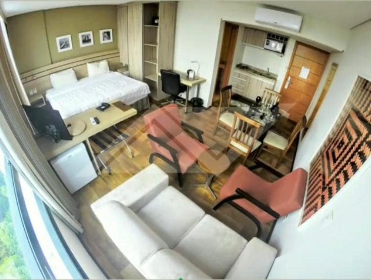 A modern living area features a spacious bed and a light-colored sofa. Two red armchairs are positioned near a wooden dining table, accompanied by a black desk chair. A kitchenette is visible in the background, with ample natural light flowing through large windows.