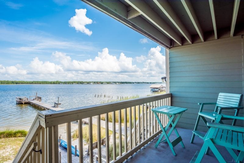 The private balcony offers serene views of the lagoon, framed by the gentle presence of clouds. Two teal chairs and a small table invite relaxation, while the peaceful water reflects light, creating an atmosphere of tranquility. A fishing pier is visible in the distance.
