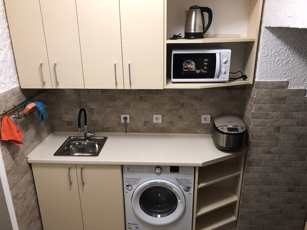 A compact kitchen area features light-colored cabinetry and a modern stainless steel kettle. A microwave sits above the countertop, while a washing machine is integrated beneath. The beige tiled wall adds texture to the space, with colorful cleaning tools hanging nearby.