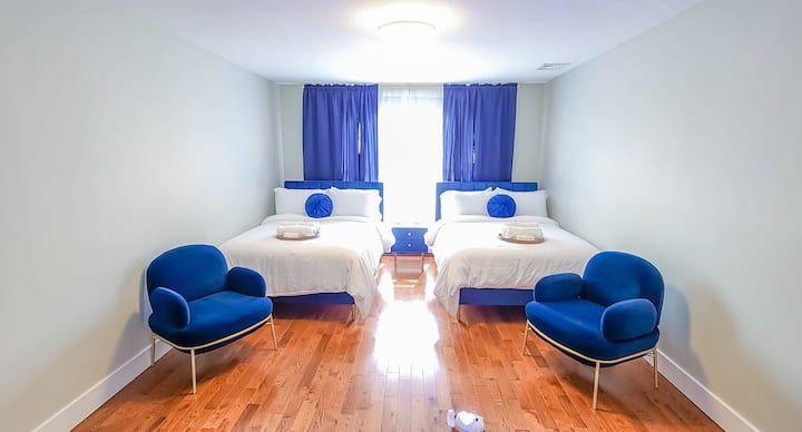 Duas camas de casal de pelúcia e detalhes azuis e brancos serenos criam um refúgio calmante, complementado por cadeiras de sofá aconchegantes para momentos tranquilos.