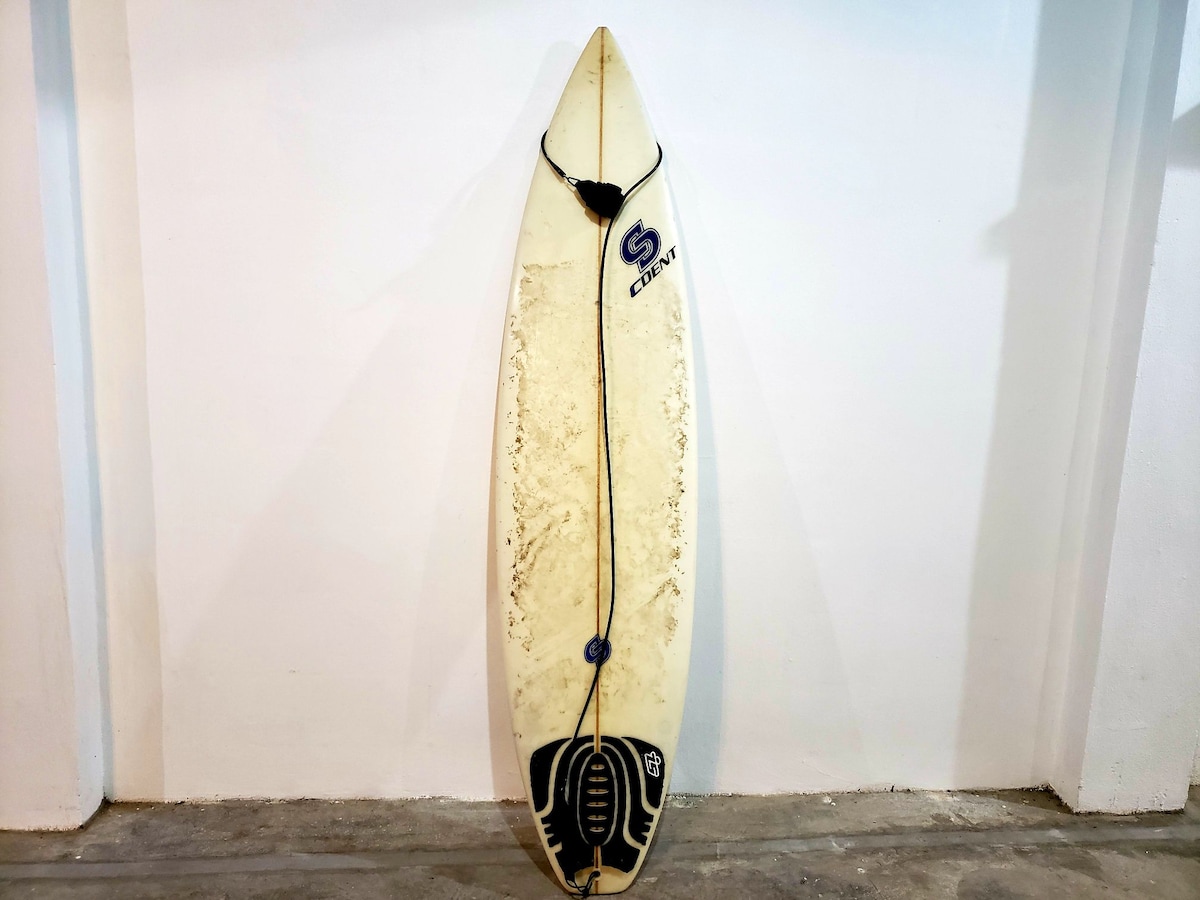 A surfboard is displayed against a plain white wall. The board shows signs of use, with visible wear and a logo near the top. A leash is attached to the tail, and the textured bottom features a prominent design that outlines its shape.