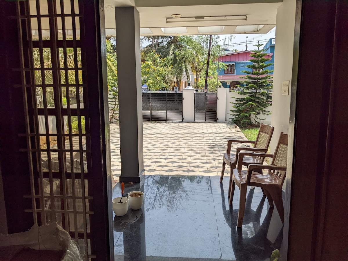 The entrance area features a polished floor leading to a spacious outdoor area. Two wooden chairs with woven backs are positioned on the left, offering a spot to relax. Greenery is visible through the entrance, adding a natural element to the view.