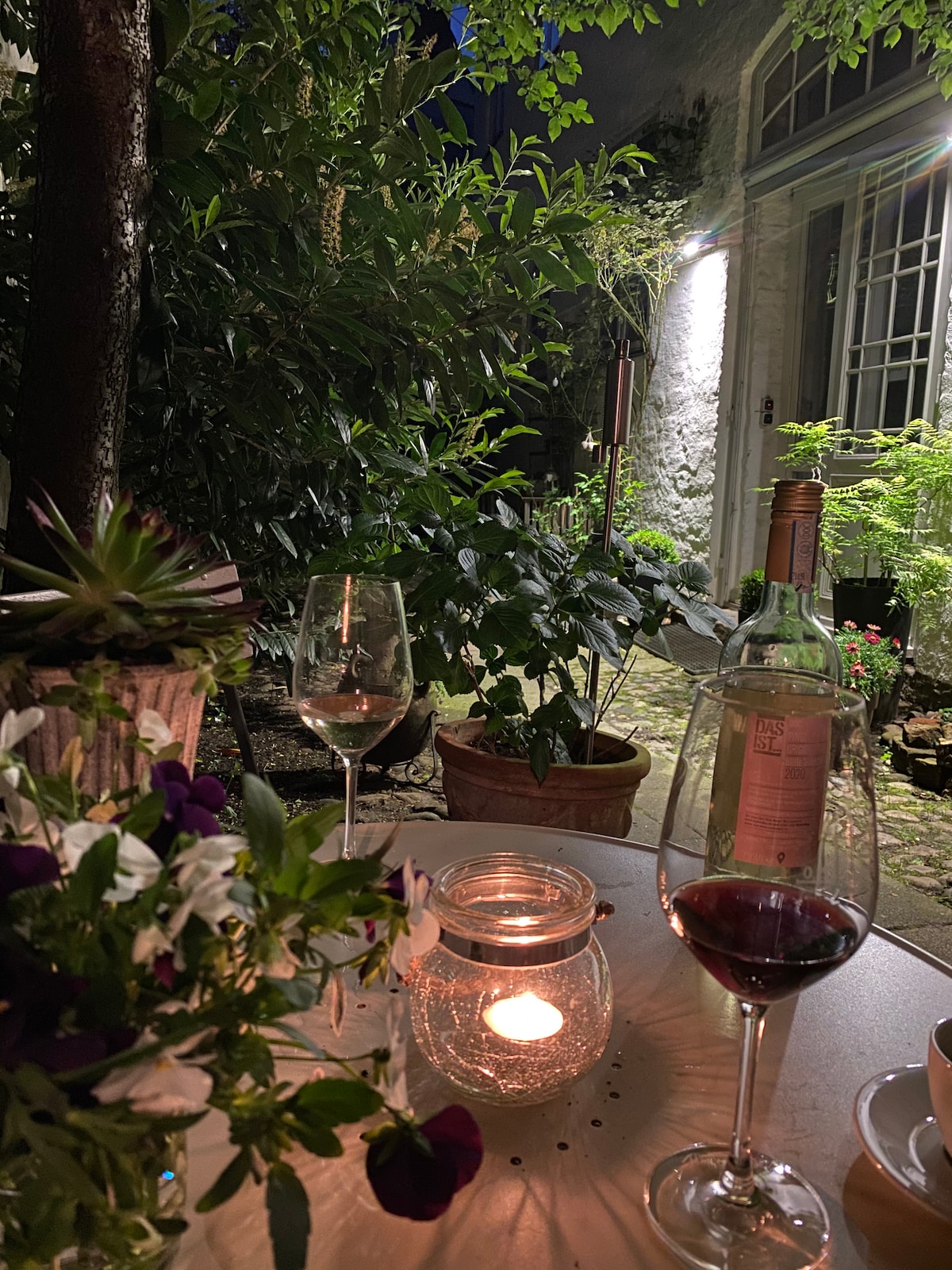 An outdoor space features a small table adorned with a bouquet of flowers and a lit candle, creating a subtle glow. Empty wine glasses sit alongside a bottle of red wine, surrounded by lush greenery and potted plants. Soft lighting highlights the tranquil atmosphere.
