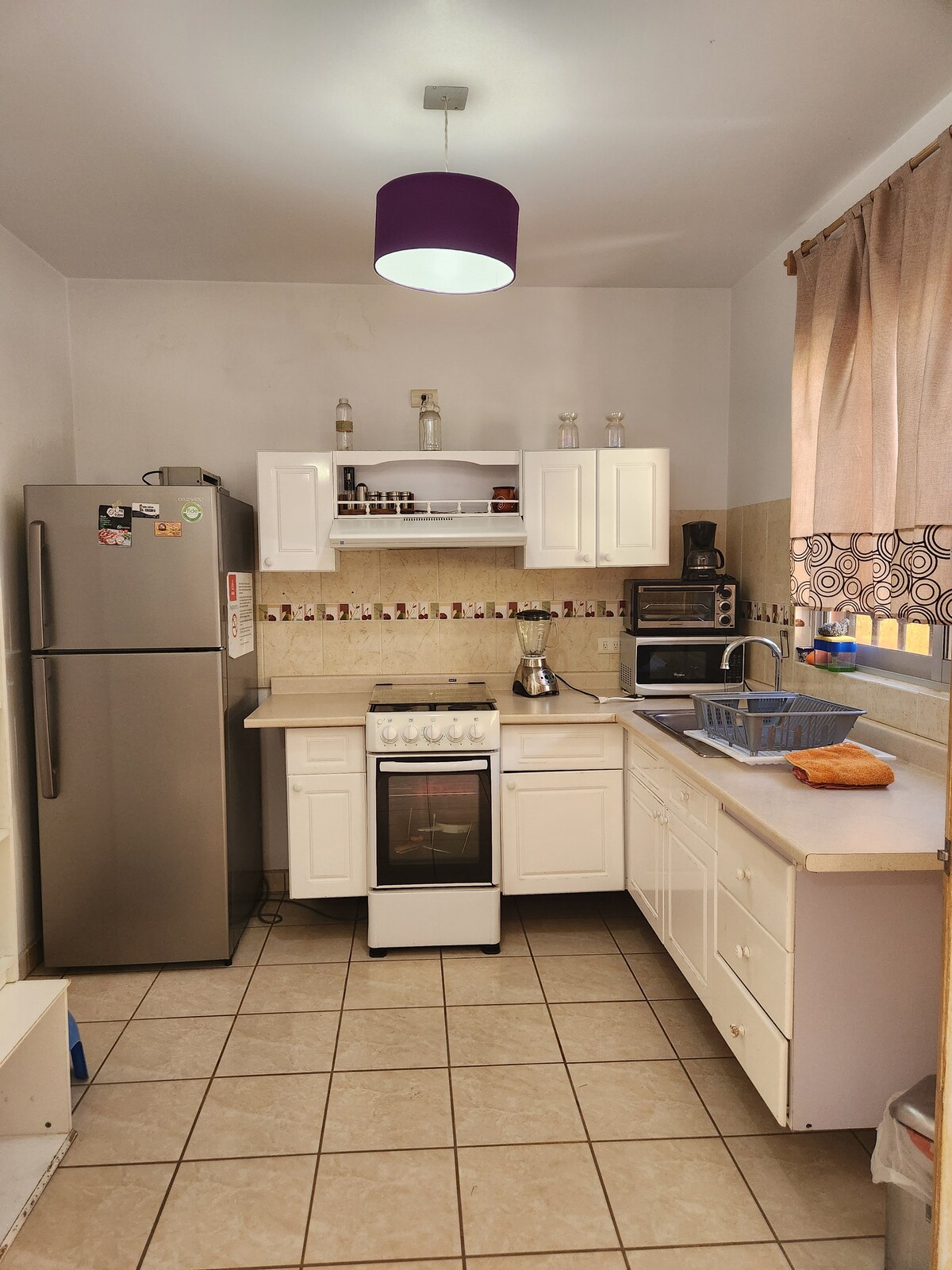 The kitchen features modern appliances, including a stainless-steel refrigerator, an oven with a stovetop, and a microwave. Light-colored cabinets offer storage space, while the counter is equipped with a sink and various implements. A window allows natural light to enter, enhancing the work area.