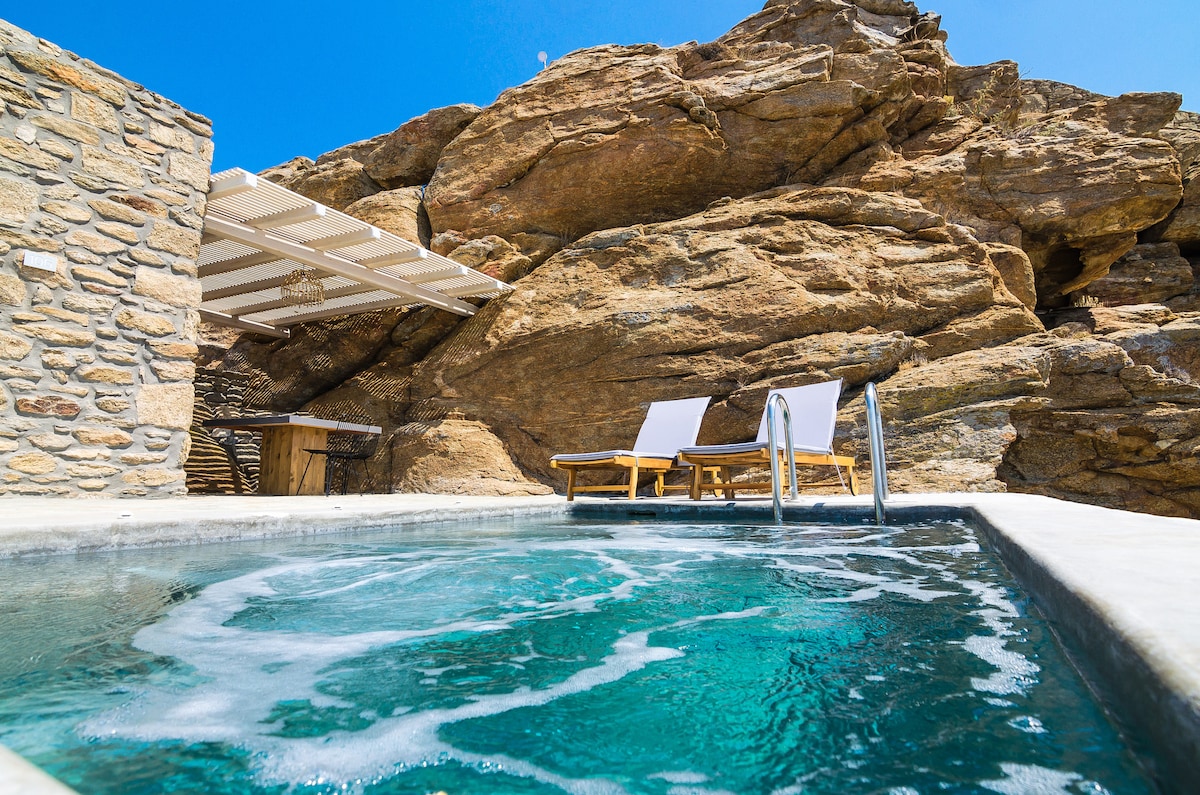 A private pool is nestled against natural rugged stones, inviting relaxation. Two loungers are positioned nearby, offering a comfortable space to unwind. A shaded area provides protection from the sun, while the clear blue sky enhances the tranquil atmosphere.