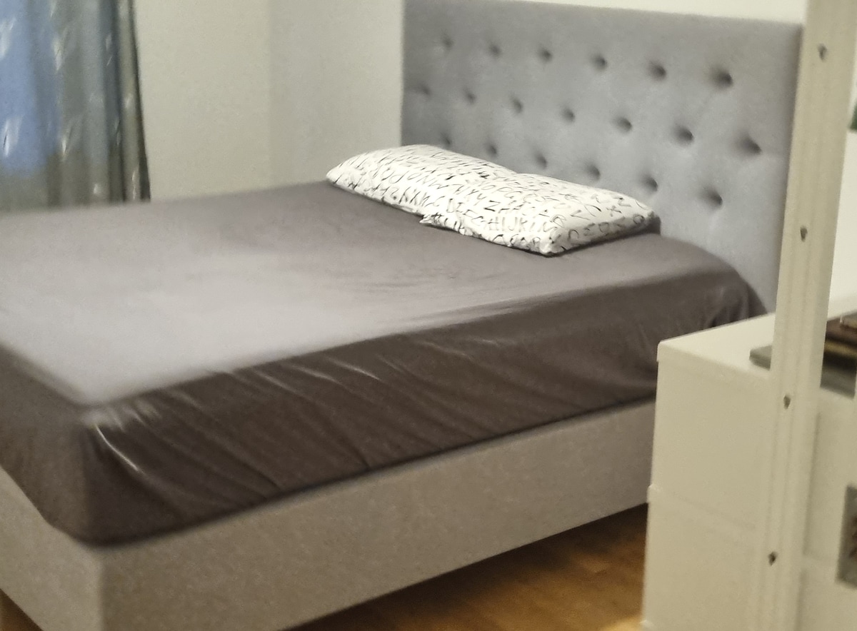 A spacious bed is presented with a tufted gray headboard and neutral bedding, complemented by a decorative pillow. Soft, wooden flooring is visible, along with a light-colored nightstand positioned nearby.