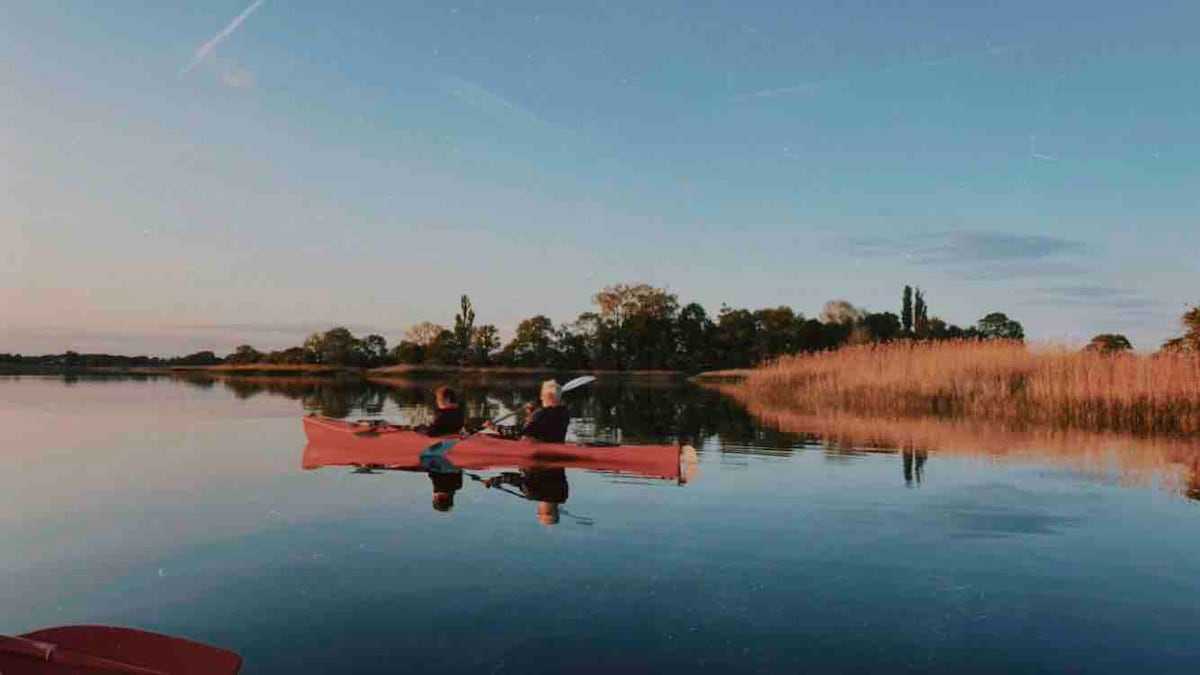 Two individuals are seated in a red kayak, gently gliding across calm waters. The surrounding landscape features tall grasses and lush trees, reflecting softly on the water's surface under a clear sky. The scene evokes a sense of peace and connection with nature.