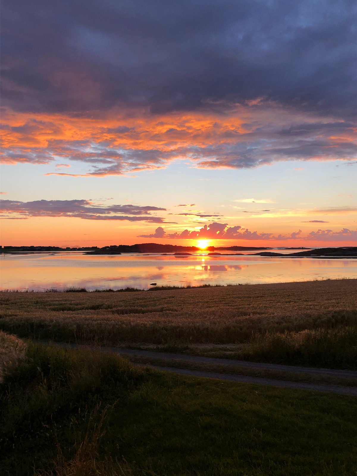 A serene sunset is captured over the calm waters, reflecting vibrant hues of orange, pink, and purple across the sky. Silhouettes of distant islands can be seen against the colorful backdrop, while golden fields creating a peaceful foreground enhance the natural beauty of the scene.