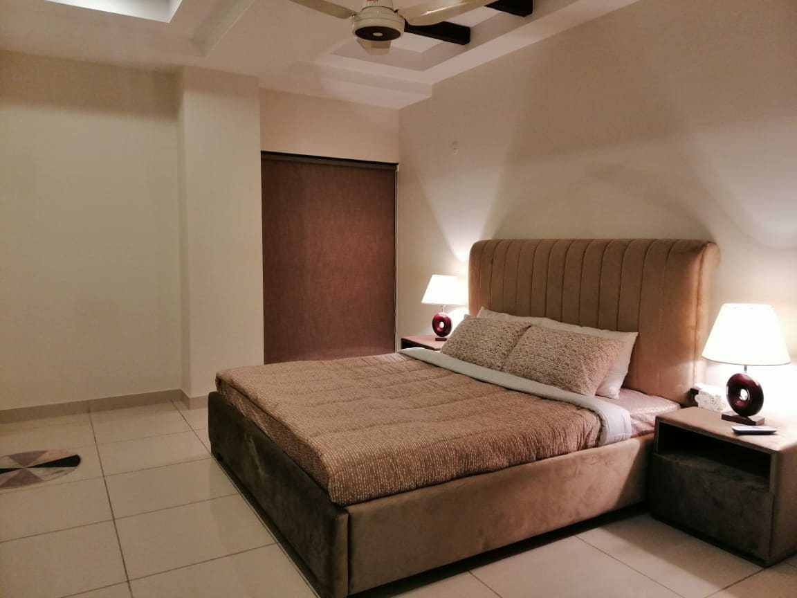 A spacious bedroom is furnished with a soft-textured bedspread on a plush bed. Two bedside lamps provide gentle lighting, while a dark curtain offers privacy. Neutral-colored walls complement the overall calming ambiance.