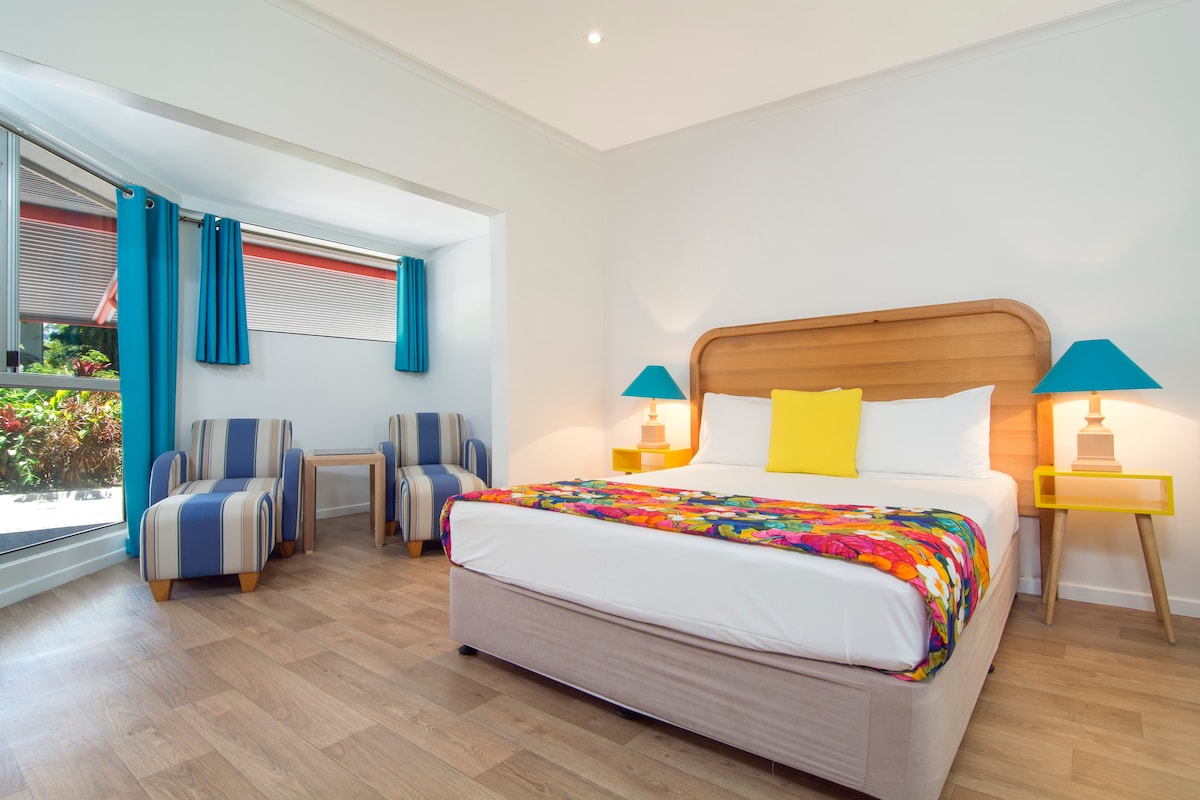 A spacious room features a comfortable bed with a colorful blanket, flanked by bedside tables with blue lamps. Two striped chairs are situated beside a small table, with natural light flooding in through large windows adorned with blue curtains.
