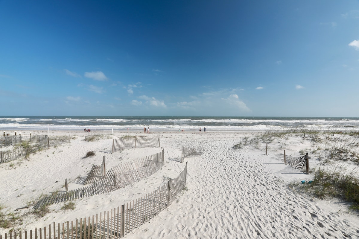The image captures a stretch of sandy beach lined with gentle dunes and wooden fencing, leading to the ocean. Waves can be seen rolling in, while a few people enjoy the shoreline under a clear blue sky with scattered clouds.