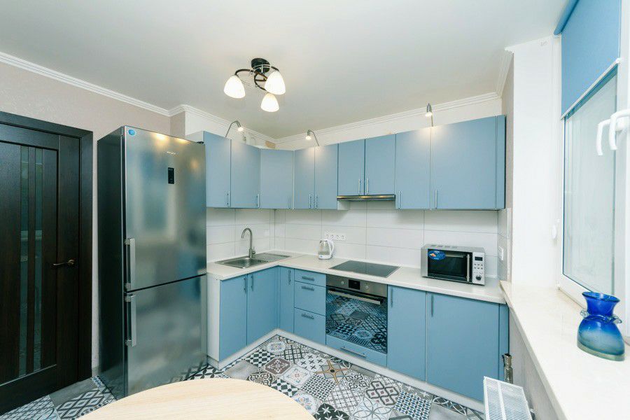 The kitchen is equipped with modern blue cabinets and stainless steel appliances, including a refrigerator and microwave. A stovetop and sink are integrated into the countertop. The floor features a patterned tile design, and light fixtures provide illumination overhead.