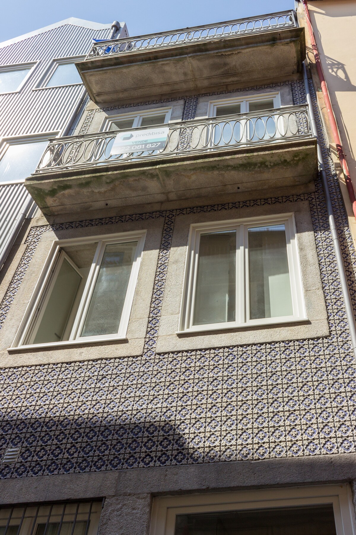 The apartment building features a decorative tiled facade in traditional Portuguese style, with two large balconies adorned by wrought iron railings. Bright, modern windows enhance the exterior, inviting natural light into the space, while the narrow street contributes to the charm of the historic center.