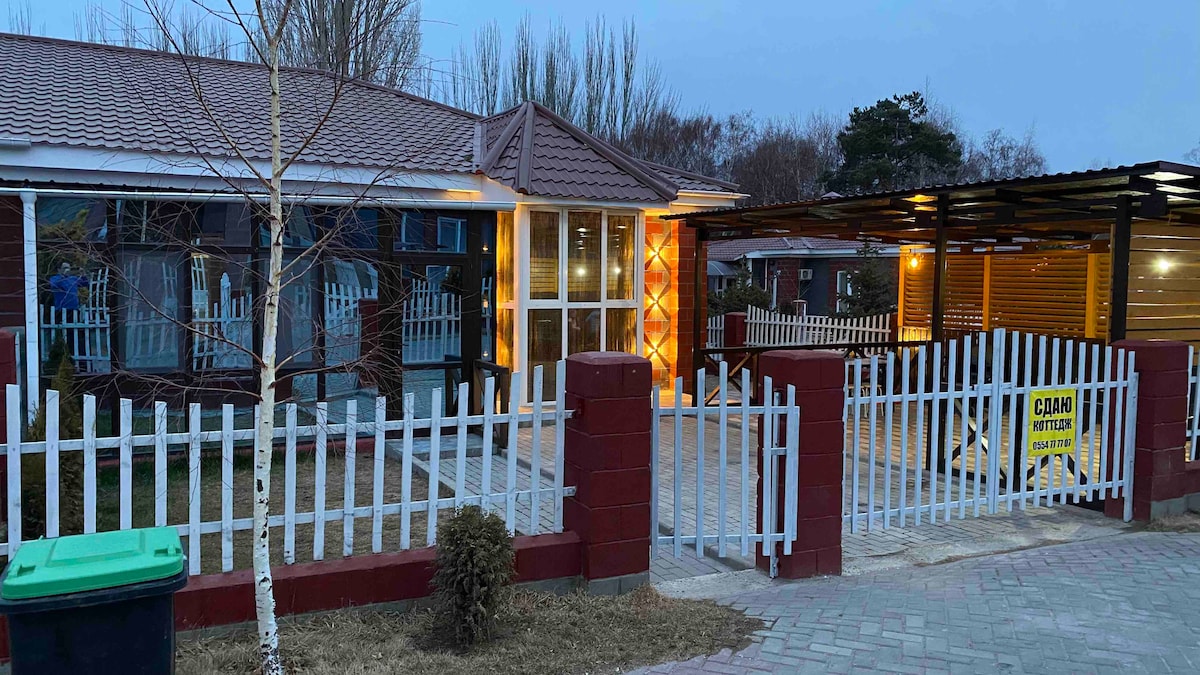 A gated entrance is visible, featuring a white picket fence that surrounds the property. Warm lighting illuminates the front porch area of a single-story structure with large windows. A covered area is shown nearby, offering additional outdoor seating options.