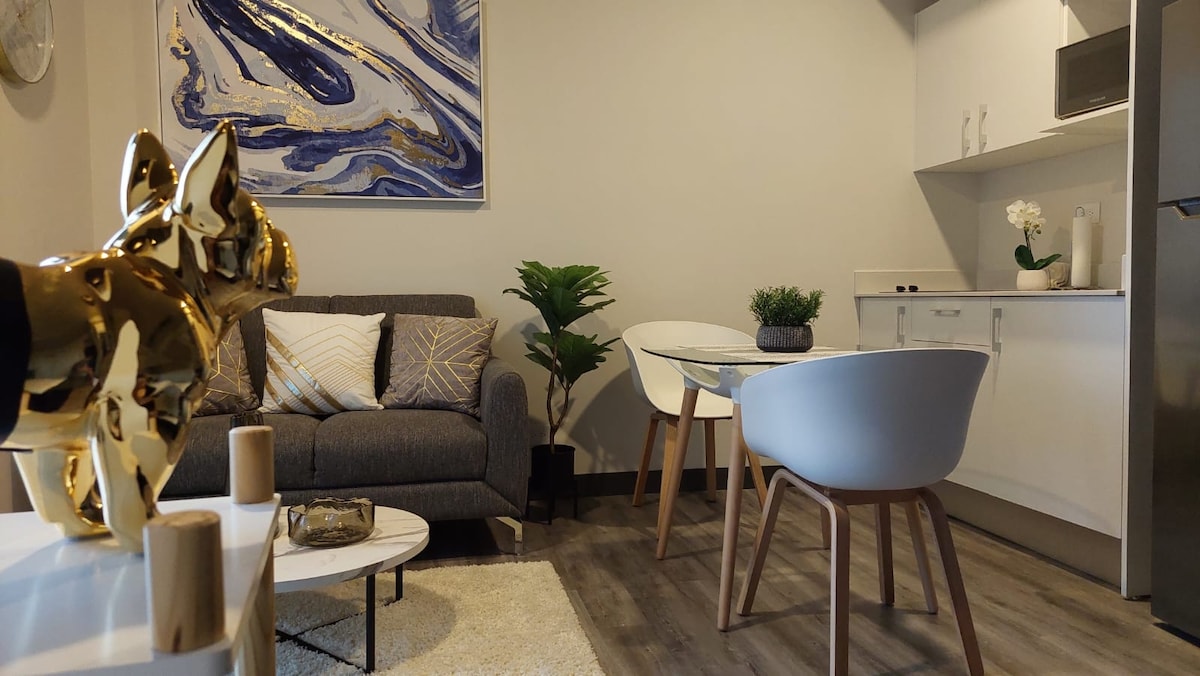 A stylish studio apartment is presented, featuring a cozy gray sofa adorned with decorative cushions. A sleek dining table with two modern chairs is positioned nearby, complemented by a small white coffee table. The kitchenette is equipped with essential appliances, enhancing the space's functionality.