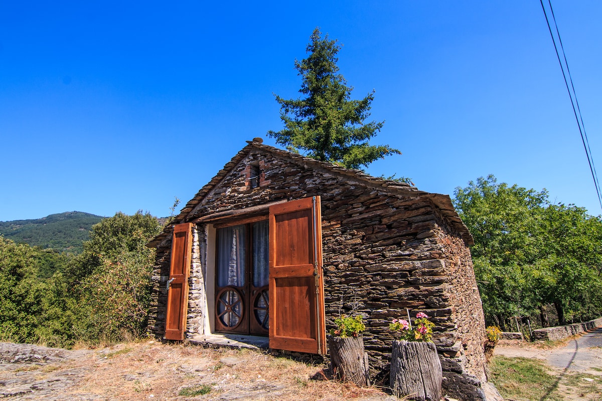 A charming stone cottage is depicted, showcasing wooden shutters on the front. Lush greenery surrounds the structure, with a clear blue sky above. The entrance features a round wooden wheel, adding character to the rustic exterior.