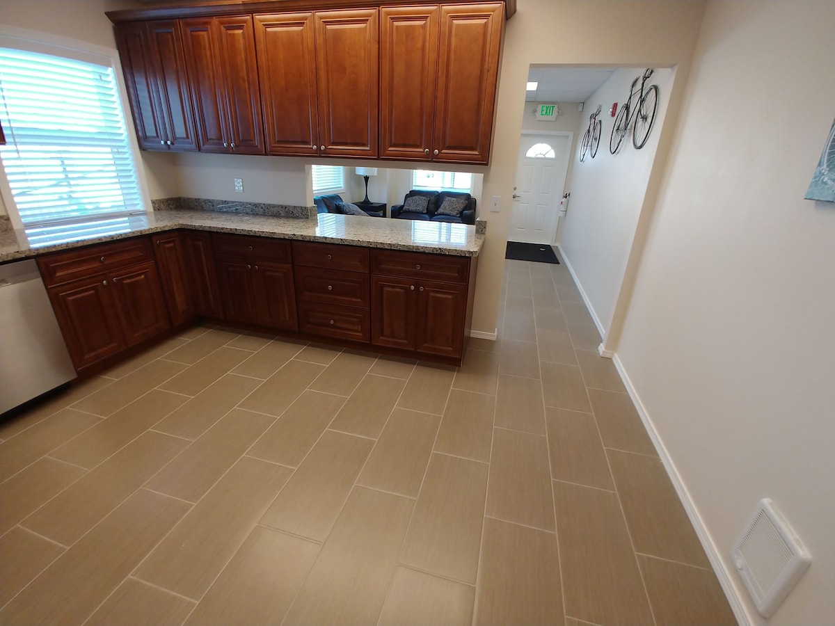 The kitchen features dark wood cabinets, granite countertops, and modern appliances. Light-colored floor tiles extend throughout the area. A doorway leads to a living room, visible in the background, while natural light filters in through a window, enhancing the welcoming environment.