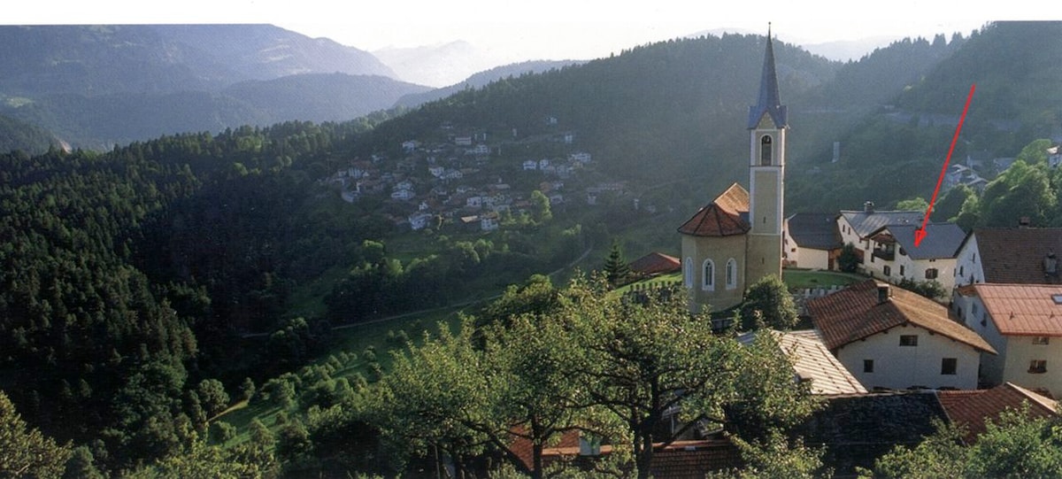 The image presents a scenic view of a mountainous landscape with a quaint village in the foreground. A church tower is visible, surrounded by traditional houses with terracotta roofs. Lush greenery stretches across the hills, creating a tranquil natural setting.