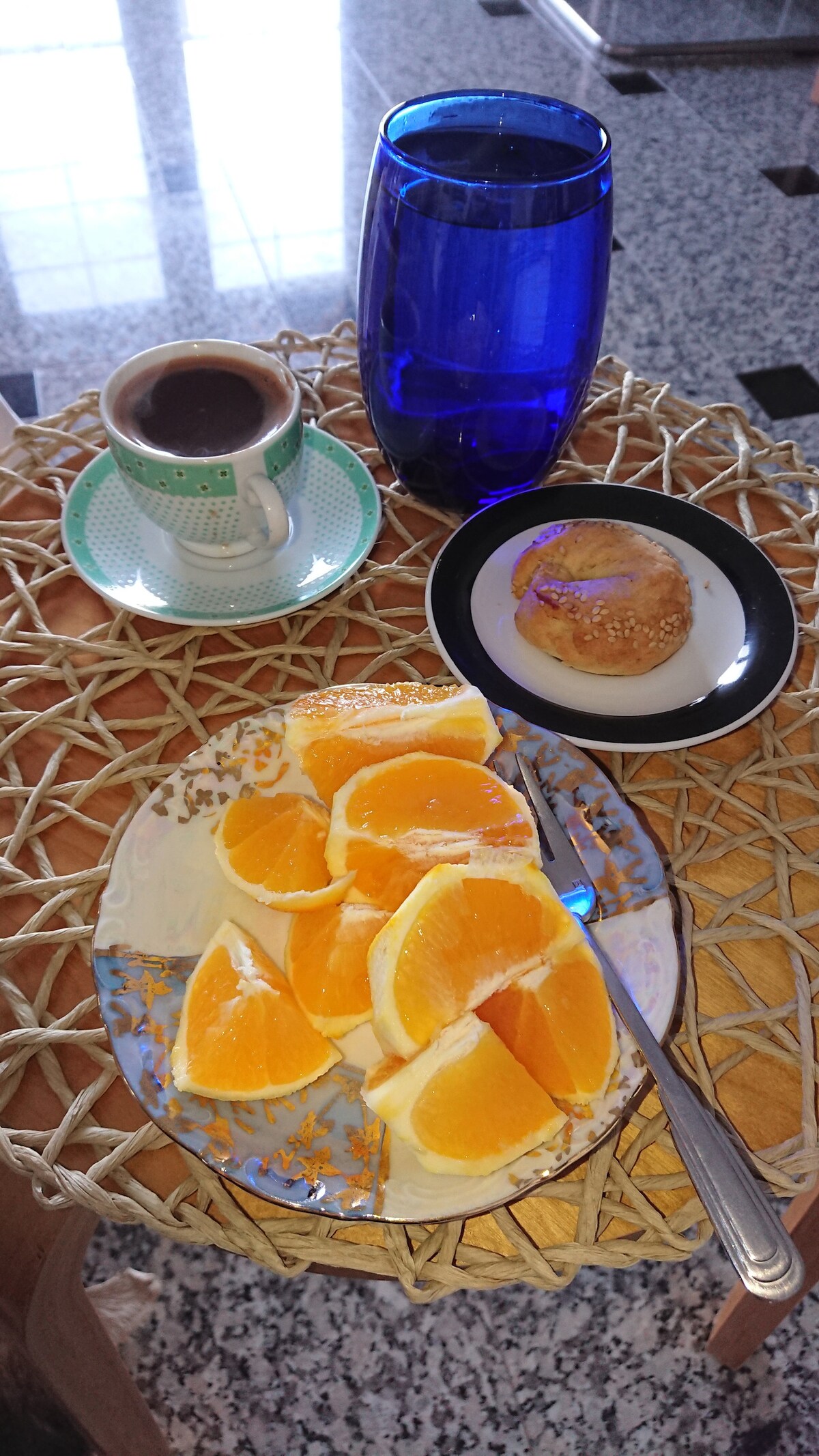 A small wooden table features a plate with neatly arranged slices of orange, alongside a black plate holding a pastry. A glass of water in a blue tumbler and a cup of coffee on a decorative saucer complete the setting. Reflections from a nearby window can be seen on the table's surface.