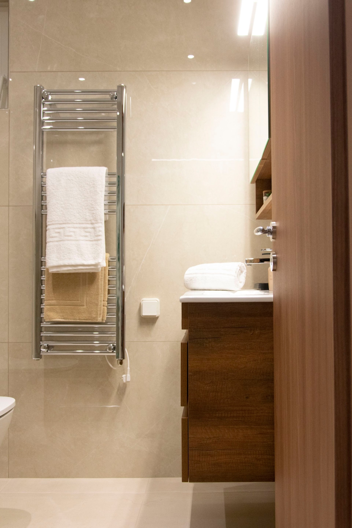 A modern bathroom is displayed, featuring a sleek towel rack holding light and dark towels. A wooden vanity with storage is visible, along with an elegant sink and a mirror above. Natural light is captured through soft lighting, enhancing the fresh ambiance.