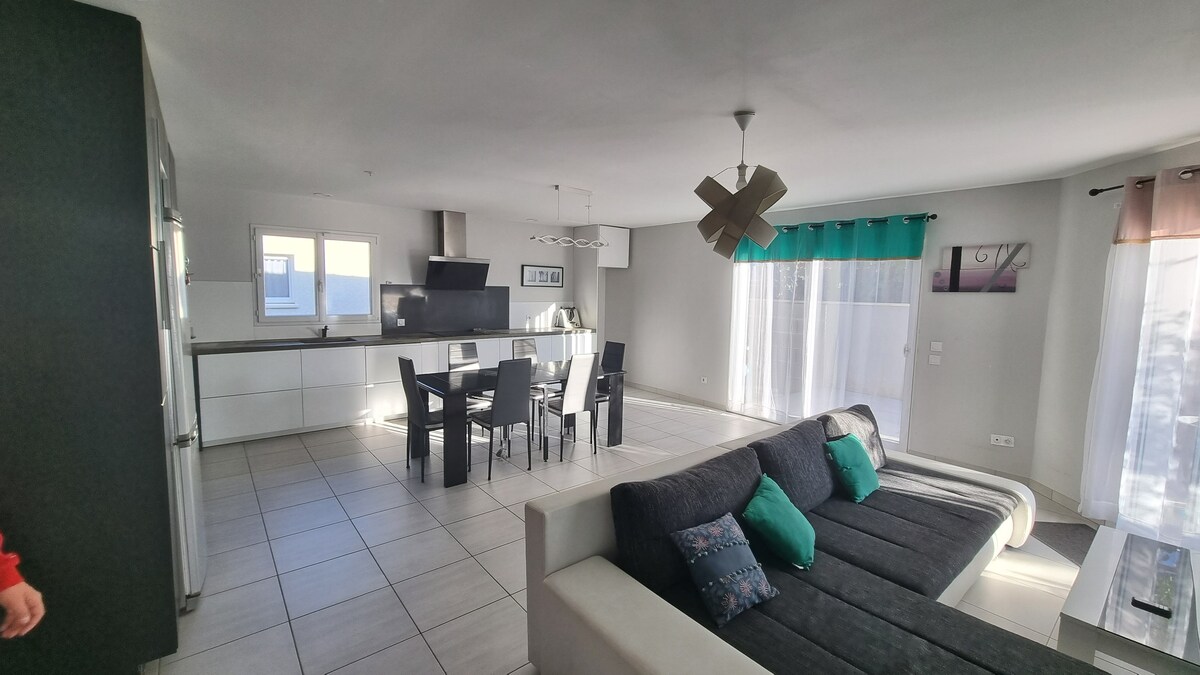 An open-concept living area features a large gray sectional sofa adorned with decorative cushions. A dining table with black chairs is positioned nearby, and a modern kitchen can be seen in the background. Natural light enters through large windows, illuminating the space.
