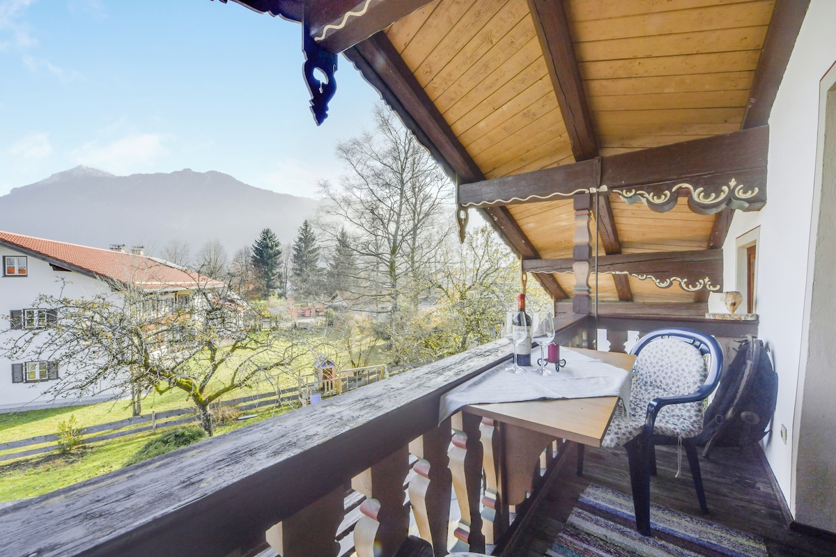 A private balcony is showcased with a table set for relaxation, featuring wine bottles and glasses. Scenic mountain views are visible beyond a lush green garden. Rustic wooden beams frame the space, providing a cozy atmosphere.