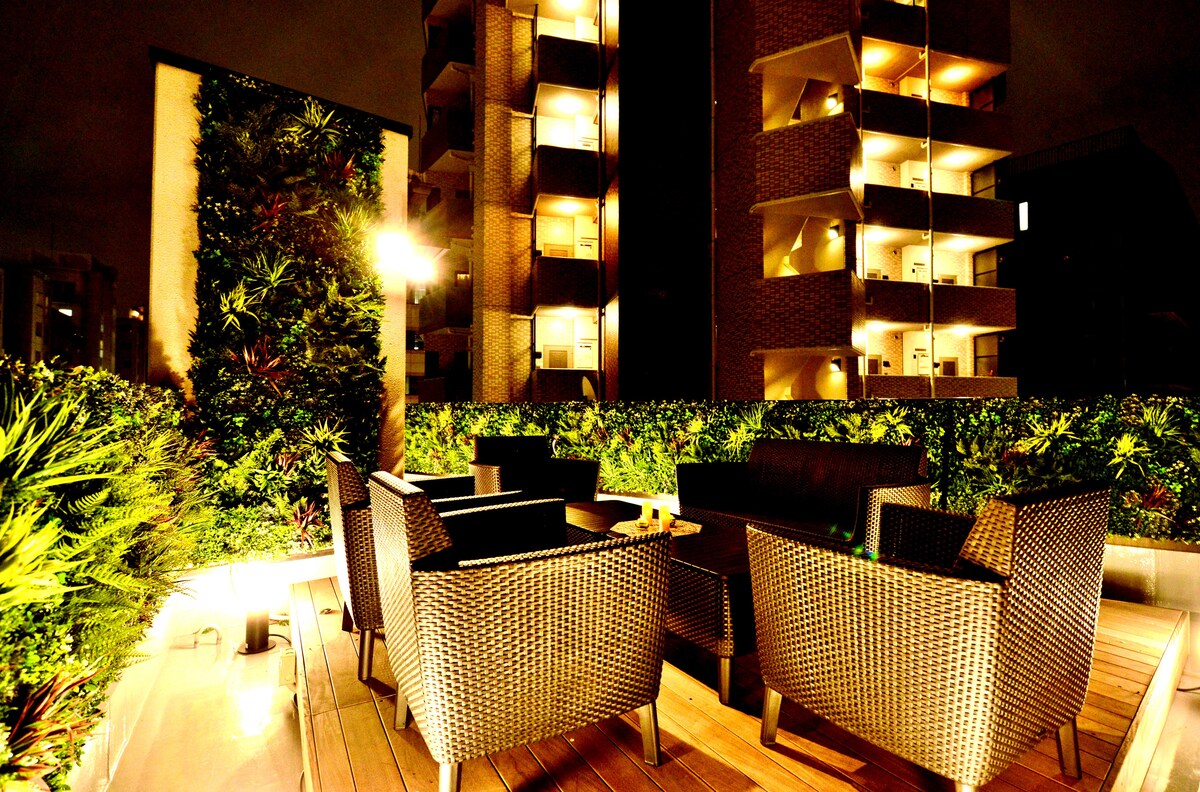 An outdoor terrace features a cozy seating arrangement with dark wicker chairs surrounding a low table. Lush greenery lines the perimeter, illuminated by warm lights, offering a serene environment at night. The backdrop includes lighted balcony structures, enhancing the urban setting.
