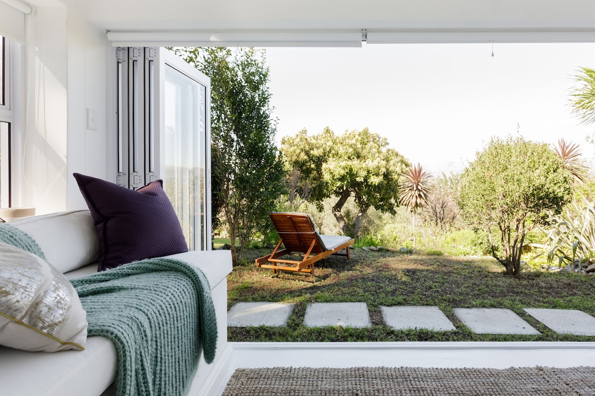 A serene view from the interior features a comfortable seating area with decorative cushions and a cozy throw. Outside, a lounge chair rests on a grassy patch, framed by lush greenery and trees leading to a pathway of stone slabs.