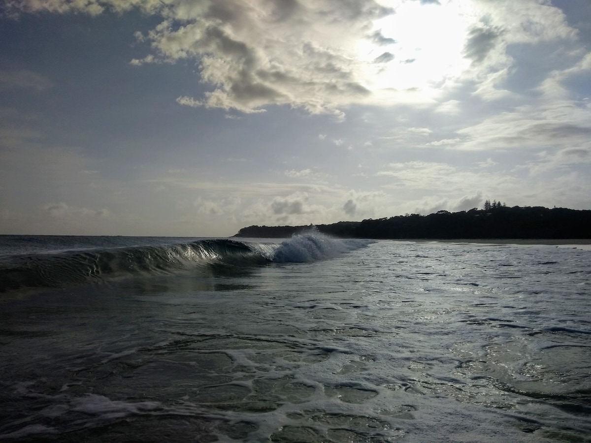Gentle waves roll onto a serene shoreline, with the sun peeking through clouds in the sky. The horizon is lined with a lush green backdrop, suggesting a coastal landscape. The surface of the water reflects soft light, creating a tranquil atmosphere.