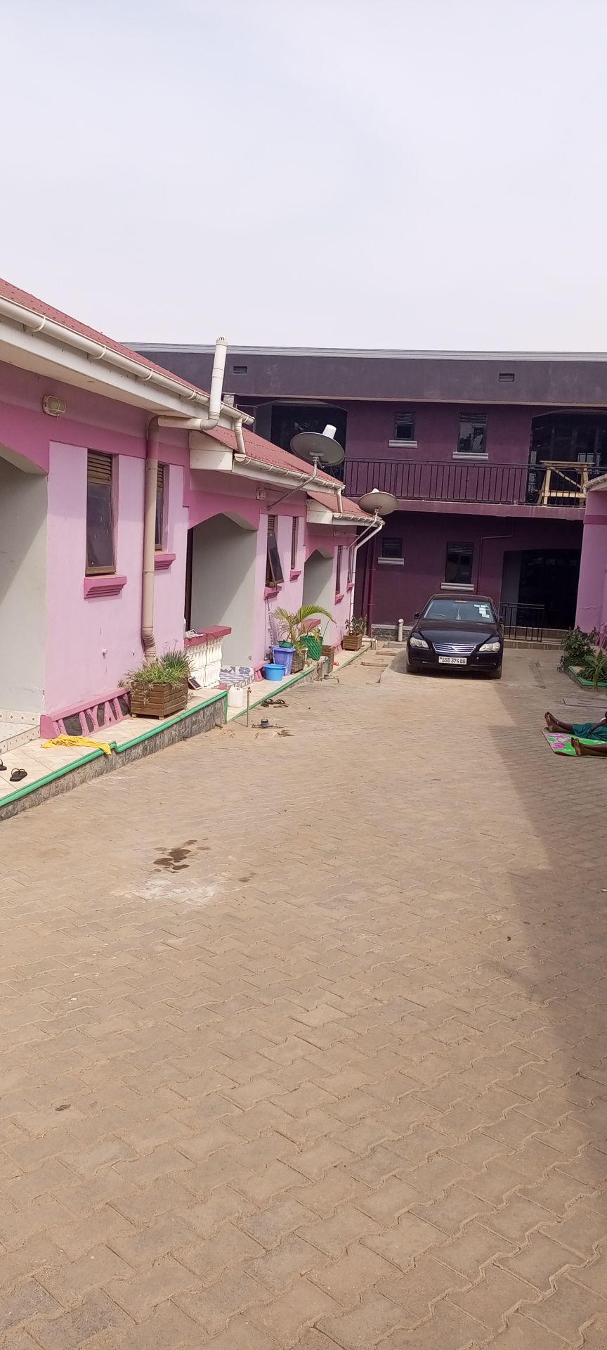 A courtyard is shown, featuring buildings with pastel pink exteriors. An open parking area is visible, containing a car. Various potted plants add greenery to the space, while the surrounding pathways are lined with colorful gardening supplies.