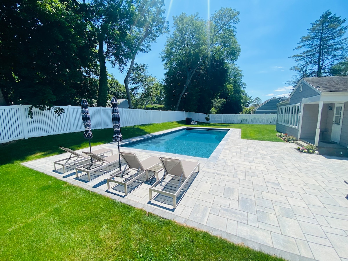 An outdoor space features a heated salt-water pool surrounded by elegant lounge chairs. Large umbrellas provide shade, while a well-maintained lawn and a privacy fence enhance the serene environment. Bright sunshine illuminates the area, creating a refreshing atmosphere for relaxation.