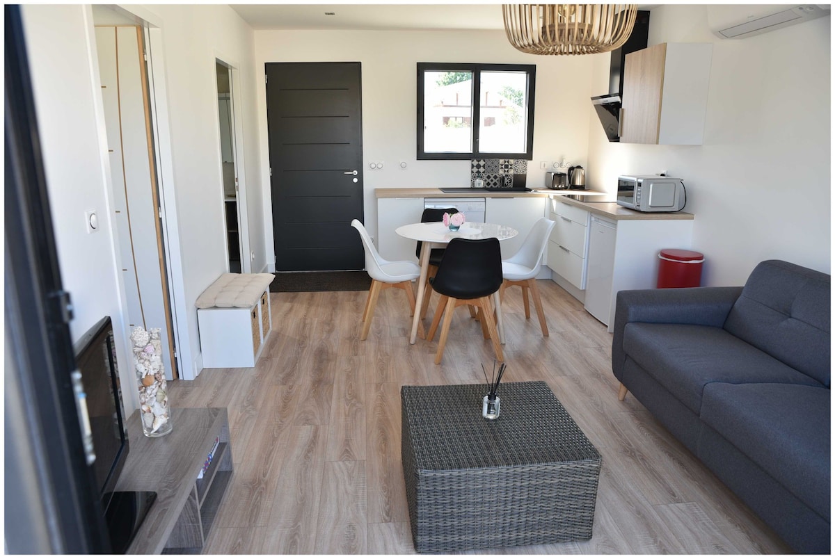 A modern living space is featured, showcasing a comfortable gray sofa and a round dining table surrounded by four chairs. A kitchenette with essential appliances is visible, complemented by light wood cabinetry. Natural light enters through large windows, enhancing the inviting atmosphere.