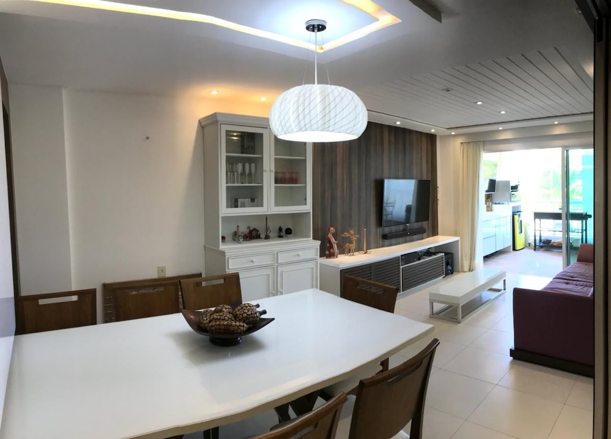 An open living and dining area is presented with a modern white dining table at the center, surrounded by wooden chairs. A stylish light fixture hangs above. A large television is mounted on a wooden accent wall, and floor-to-ceiling windows provide natural light and views of the outdoor space.