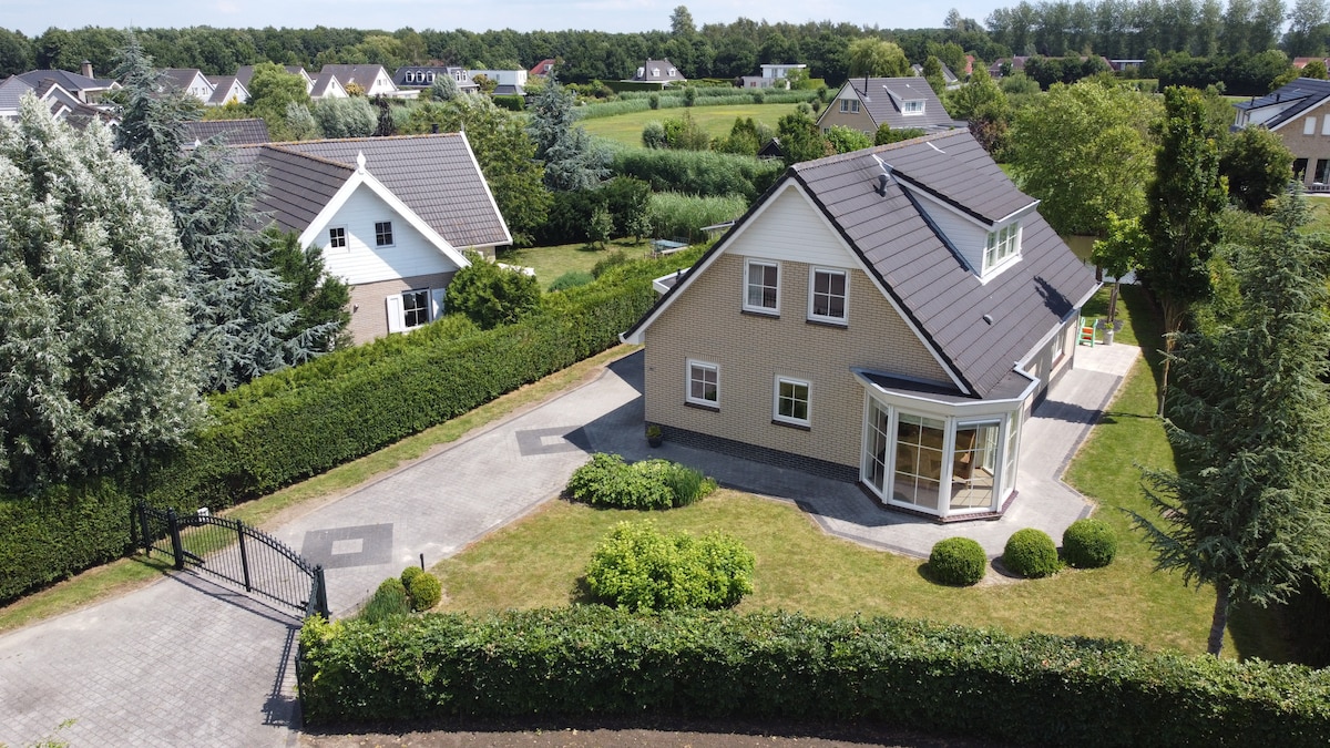 A spacious, light-colored two-story house is shown from an elevated view. Surrounded by well-maintained gardens, the property features a paved driveway leading to a welcoming entrance. Lush greenery and trees outline the yard, enhancing the tranquil setting. The picturesque landscape showcases neighboring houses and open fields.