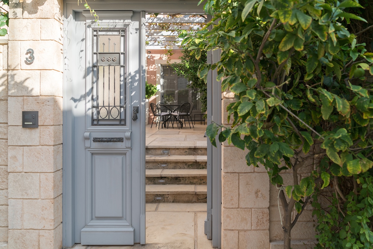 A grey door with decorative details is framed by lush greenery, leading to a charming patio area. Stone steps ascend to a spacious outdoor seating arrangement, where black chairs and a table are partially visible, surrounded by creeping vines and sunlight.