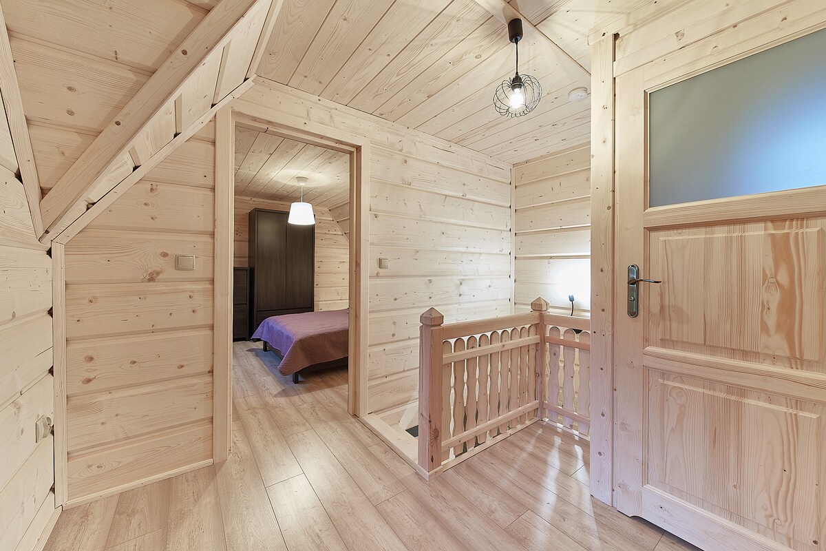 A wooden hallway features natural light filtering through a window, highlighting the light-colored wood paneling and flooring. A decorative railing is situated to one side, while a doorway leads to a spacious bedroom beyond, indicated by a soft-hued blanket on the bed.