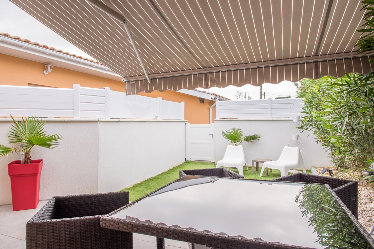 A covered terrace area features a glass-top table surrounded by wicker chairs. Sun loungers are positioned against a wall with greenery and a potted palm. A privacy fence is visible, creating an intimate outdoor space.