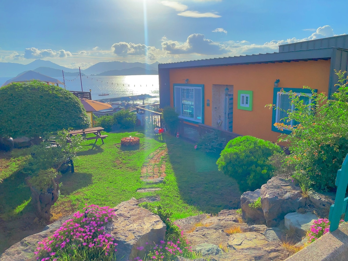 A vibrant garden is displayed, featuring well-maintained greenery and flowering plants. A cozy orange structure with blue accents is visible in the background, along with pathways made of natural stone. The scene is illuminated by soft sunlight, with views of distant hills and water.