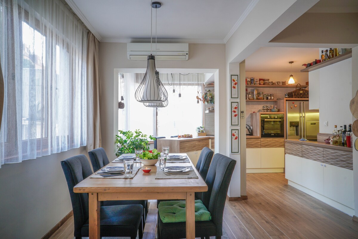 A dining area features a wooden table surrounded by dark blue chairs, set for a meal with glassware and a small bowl. Natural light enters through large windows, highlighting greenery outside. In the background, a modern kitchen is visible, showcasing cabinets and appliances.
