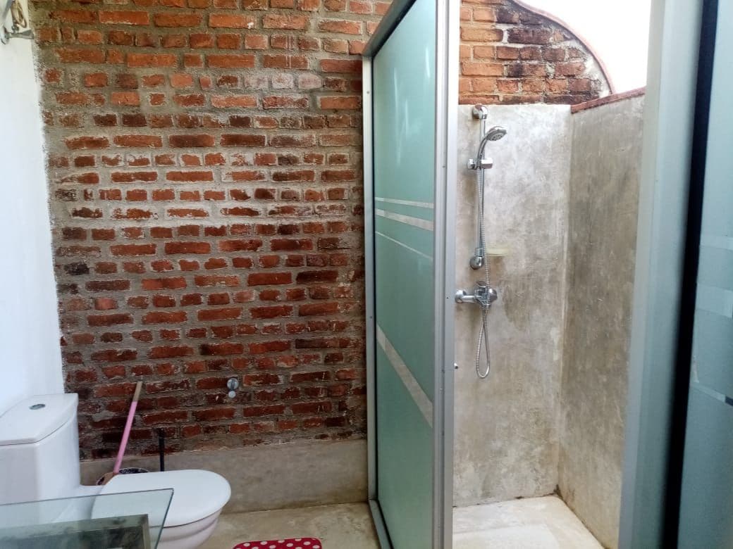 A modern bathroom features a walk-in shower enclosed by frosted glass, with sleek metal fixtures. Exposed brick walls add a rustic charm to the space, complemented by a minimalist white toilet. The floor showcases neutral tiles, providing a clean and functional environment.