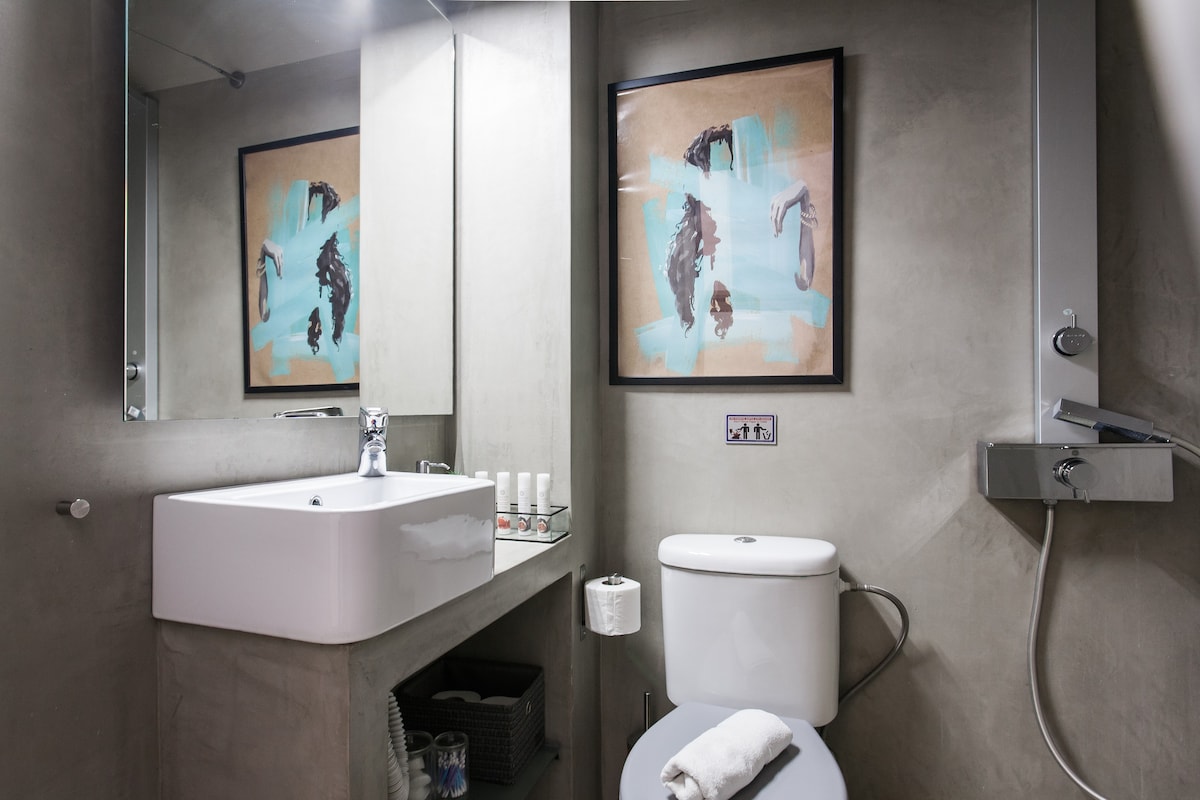 The modern bathroom features a sleek concrete wall and a large mirror above a minimalist sink. A stylish piece of wall art adds a splash of color. Essential toiletries are neatly arranged on the counter, while a roll of toilet paper is positioned beside the clean, compact toilet.