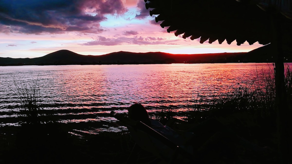 The image captures a serene sunset over Lake Titicaca, with vibrant hues of orange, pink, and purple reflected on the water. Silhouetted mountains create a tranquil backdrop, while the foreground features gentle waves lapping at the shore, adding to the peaceful ambiance.